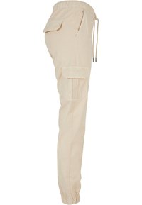 Pantalon cargo beige avec taille élastique, cordon de serrage, poches latérales, poche à rabat sur la cuisse et poignets élastiques aux chevilles.