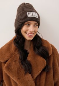 Lachende vrouw met lang donker haar die een bruine gebreide muts draagt met het label "BIZUU" en een grote bruine nepbontjas, tegen een effen achtergrond.