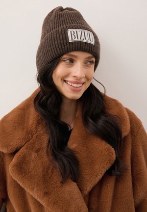 Lachende vrouw met lang donker haar die een bruine gebreide muts draagt met het label "BIZUU" en een grote bruine nepbontjas, tegen een effen achtergrond.