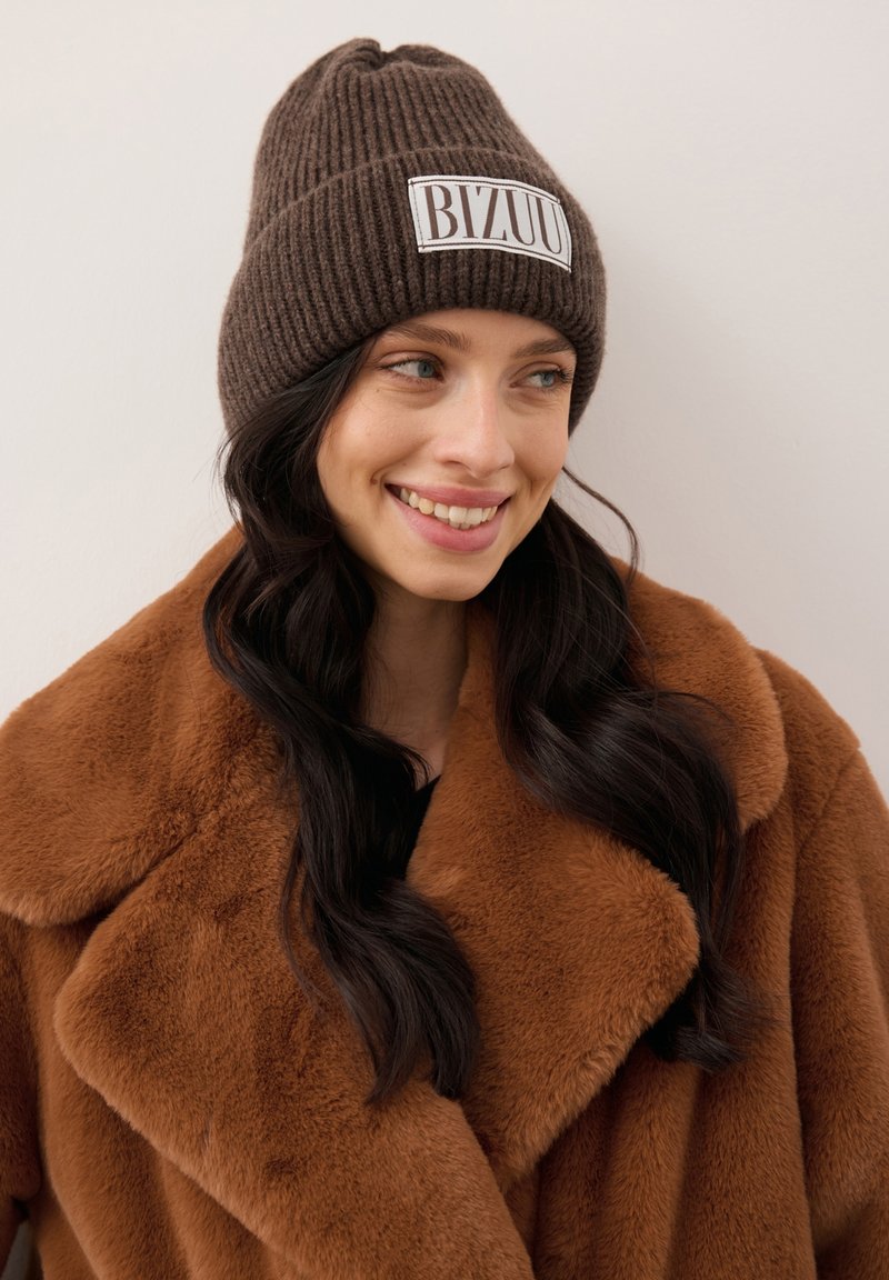 Lachende vrouw met lang donker haar die een bruine gebreide muts draagt met het label "BIZUU" en een grote bruine nepbontjas, tegen een effen achtergrond.