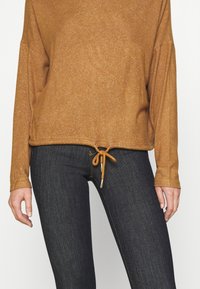Maglia marrone a maniche lunghe con orlo regolabile e una texture sottile, abbinata a jeans di denim scuro con una vestibilità classica e cuciture fini.