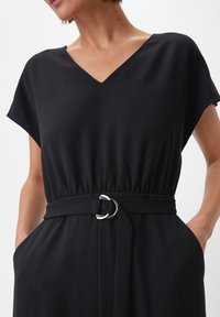 Czarna sukienka z krótkimi rękawami i dekoltem w kształcie litery V. Posiada elastyczny pas oraz materiałowy pasek z srebrną klamrą w kształcie pierścienia. Gładka tekstura.