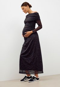 Robe maxi à épaules dénudées en tissu sombre à motifs, avec fronces, manches longues transparentes, portée avec des baskets noires et blanches.