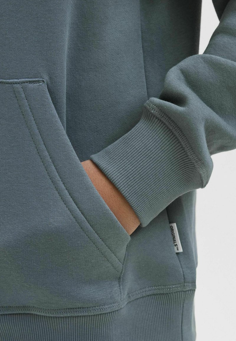 Grön sweatshirt med känguruficka. Har ribbade mudd och nederkant, mjukt tyg och ett litet märke med "ORIGINALS" på sidan.