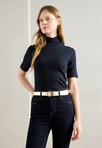 Chemise à col roulé côtelé marine à manches courtes, associée à un jean en denim sombre taille haute et une ceinture blanche avec une boucle circulaire.