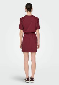Bordeaux kort jurkje met elastische taille, korte mouwen en witte zijstripes. Gemaakt van een zachte stof, met een relaxte pasvorm.