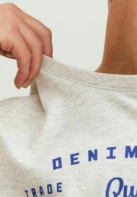 Lysegrå T-shirt med blå tekst, der har en rund halsudskæring og en blød, glat tekstur, holdt af en hånd ved kraven.