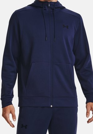 Mann trägt einen marineblauen Kapuzenpullover mit Reißverschluss und passende Hosen mit schwarzen Under Armour-Logos auf Brust und Oberschenkel.