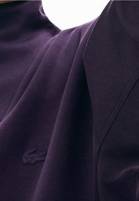 Prenda de algodón morado con cuello alto que presenta un acabado texturizado y un pequeño logo de cocodrilo en tono similar. Los detalles de las costuras son sutilmente visibles.