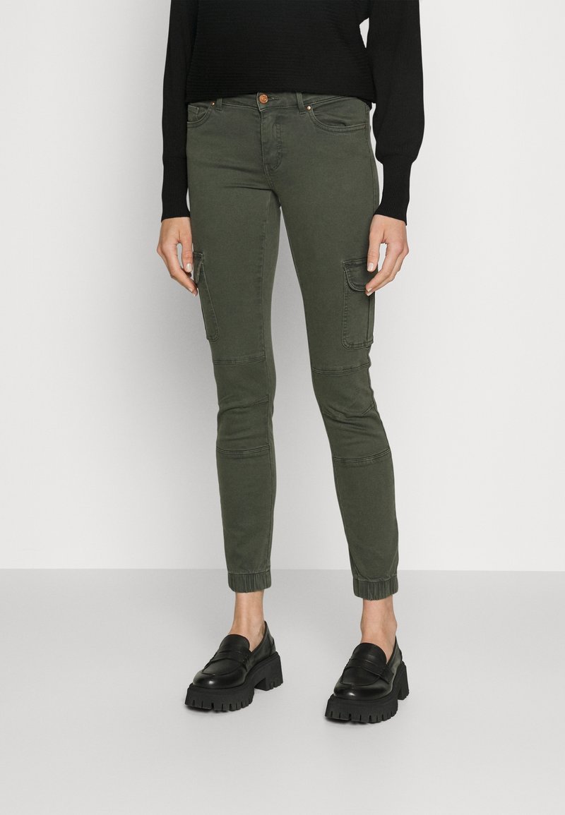 Femme portant un pantalon cargo vert olive avec des poignets élastiques, un pull noir à manches longues, et des mocassins à plateforme noirs et épais.
