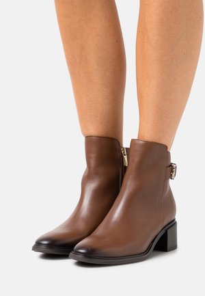 Brązowe skórzane botki za kostkę z delikatnym połyskiem, z bocznym zamkiem błyskawicznym oraz metalowym zapięciem na kostce. Projekt na słupkowym obcasie.
