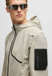 Light grey lightweight jacket with geometric pattern, adjustable hood, black accents, and zippered pocket on the left sleeve.