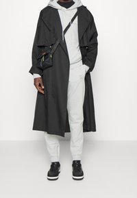 Abrigo de trench negro con frente abierto, sudadera gris con capucha y pantalones de chándal gris claro. Zapatillas negras con suelas blancas. Pequeña bolsa cruzada negra.