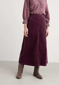 Garšīgs, tumši vijolītes krāsas korduroja svārks ar taisnu siluetu, kuram ir teksturēta virsma un augsta vidukļa līnija, apvienots ar brūnām potīšu zālēm.