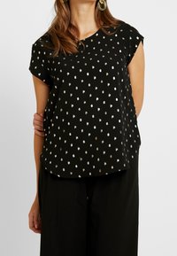 Top corto nero a maniche corte con motivo a pois metallici oro e argento, scollo rotondo e tessuto morbido. Abbinato a pantaloni neri larghi.