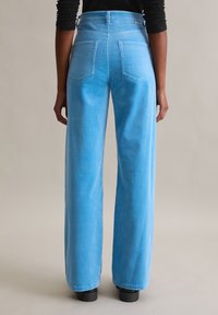 Pantalon large en velours côtelé bleu clair, taille haute, avec deux poches arrière et des rainures verticales subtiles dans la texture du tissu.