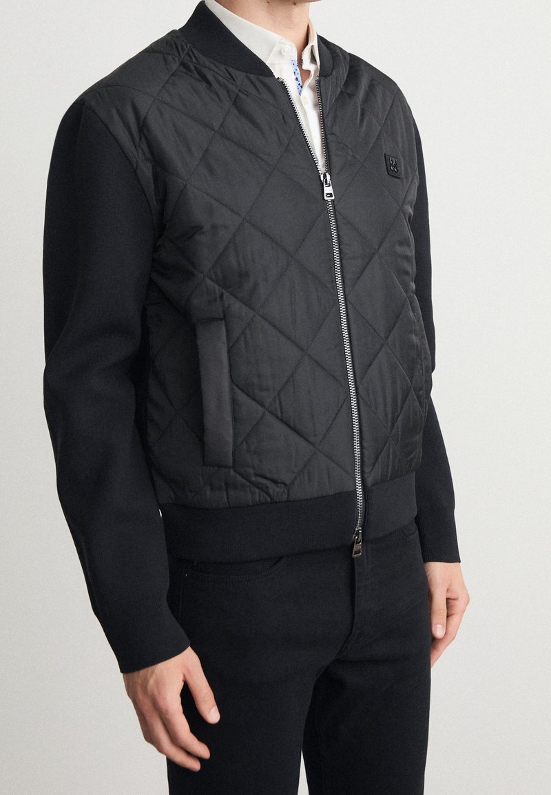 Blouson aviateur matelassé noir avec fermeture éclair à l'avant, poignets côtelés et une poche. Tissu mat à texture lisse avec une coupe ajustée.