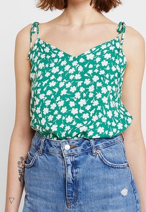 Kobieta nosząca zielony top na cienkich ramiączkach z białym kwiatowym wzorem oraz niebieską dżinsową spódnicę z wysokim stanem, pokazująca tatuaże na ramionach.