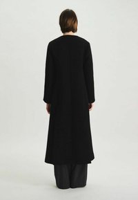 Manteau noir long au design épuré, en tissu texturé, avec de longues manches, porté par-dessus un pantalon à rayures fines. La vue de dos met en avant une silhouette simple.