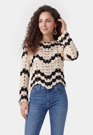 Kvinde med krøllet hår iført sort og cremefarvet hæklet sweater og blå jeans, der rører ved håret og kigger let til siden.