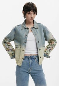 Jeansjacke mit bestickten Blumenmotiven an den Ärmeln, Knopfschluss, hellblaue Waschung und zwei Fronttaschen. Über einem weißen Oberteil getragen.
