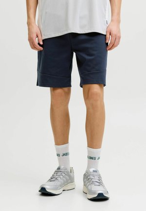 Muškarac u mornarskoplavim kratkim hlačama do koljena, bijelim čarapama Jack & Jones i srebrnim tenisicama, stoji na jednostavnoj svijetloj pozadini.