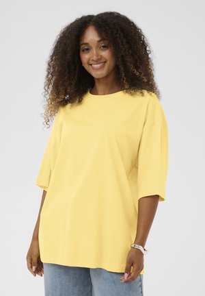 Usmievavá žena s kučeravými vlasmi, oblečená v voľnej žltej košeli s krátkym rukávom a svetlomodrých džínsach na jednofarebnom sivom pozadí.