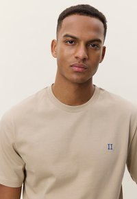 Beige bomull t-skjorte med rund hals, med en blå "II"-logo på brystet. Korte ermer og glatt tekstur.