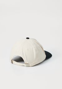 Casquette de baseball en tissu coton écru avec une visière noire. Elle présente un bouton noir sur le dessus et une fermeture par sangle à l'arrière.
