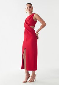 Red sleeveless dress with a twisted front design, cut-out details at the waist, and a side slit. Paired with strappy gold heels.