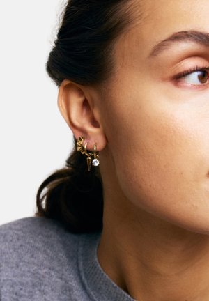 Orecchini color oro con una varietà di forme geometriche, tra cui una stella, un picco e un elemento in cristallo, appesi a un design ad anello.