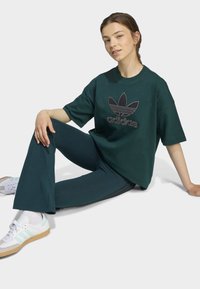 Temno zelena oversize majica z črnim logotipom Adidas, v kombinaciji s temno zelenimi širokimi hlačami in belimi supergami z mentolovimi črtami.