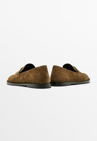 Mocassini in suede marrone con un design semplice, suole nere piatte e dettagli cuciti sulla parte superiore. Texture liscia con uno stile minimale.