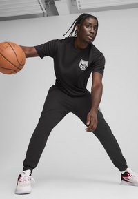 Juodas sportinis komplektas, kuriame yra trumpomis rankovėmis marškinėliai su balta Puma logo, siaurėjantys kelnių ir baltos batai su raudonais akcentais, laikantys krepšinį.
