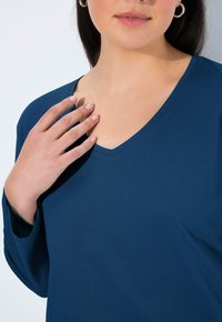 Marineblauwe V-hals top van een zachte, gestructureerde stof. Lange mouwen en een ontspannen pasvorm. Een hand rust voorzichtig op de halslijn, met zichtbaar natuurlijke nagels.