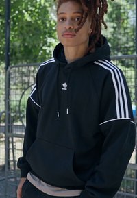 Felpa nera con accenti bianchi e design a tre strisce, dotata di tasca a marsupio e cappuccio regolabile con cordino. Realizzata in tessuto morbido.