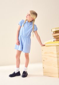 Fată tânără în salopetă albastră cu model gingham, cu cozi împletite, se sprijină de o masă de lemn pe care sunt așezate cărți, privind în sus spre stânga.