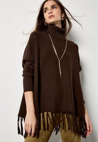 Brązowy dziany poncho z wysokim kołnierzem, z frędzlami na dolnym końcu i prążkowanymi rękawami. W połączeniu z oliwkowymi spodniami.
