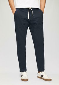 Marine stretch katoenen joggers met elastische tailleband, trekkoord, zijzakken en taps toelopende benen. Draagt een paar witte en tan sneakers.