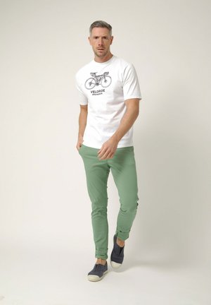 Hombre caminando hacia adelante con una camiseta blanca con un gráfico de bicicleta y pantalones verdes, sobre un fondo claro y sencillo.