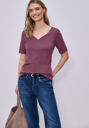 Uśmiechnięta kobieta w krótkim różowym koszulce z dekoltem w serek, niebieskich dżinsach z czarnym paskiem, trzymająca brązową torbę na ramię, stojąca na tle jednolitego jasnego tła.