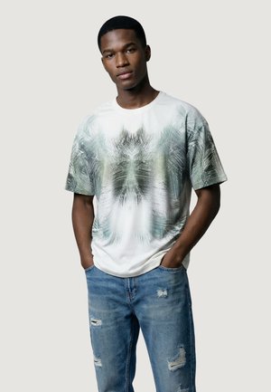 Młody mężczyzna z krótkimi włosami, ubrany w białą koszulkę z zielonymi abstrakcyjnymi wzorami i podniszczone niebieskie dżinsy, ręce w kieszeniach, neutralne tło.