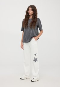 Szara, oversize'owa koszulka z okrągłym dekoltem, zestawiona z białymi spodniami o szerokiej nogawce z ciemnymi wzorami gwiazd po lewej stronie. Casuale buty sportowe.