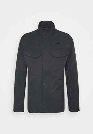 Schwarze Nike-Jacke mit Stehkragen, zwei Brusttaschen und Druckknopfverschluss vorne. Sie zeichnet sich durch ein glattes, synthetisches Material und ein Logo-Detail auf der Brust aus.