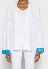Bílý otevřený pletený cardigan s texturovaným vzorem listů, zdobený modrými akcenty na manžetách s označením "AIX." Nosený přes bílý košili s V-výstřihem.