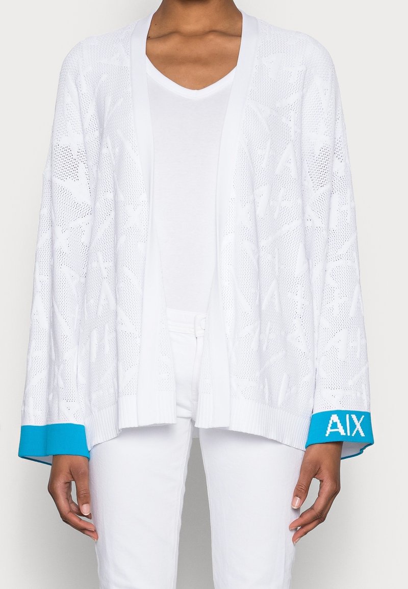 Bílý otevřený pletený cardigan s texturovaným vzorem listů, zdobený modrými akcenty na manžetách s označením "AIX." Nosený přes bílý košili s V-výstřihem.