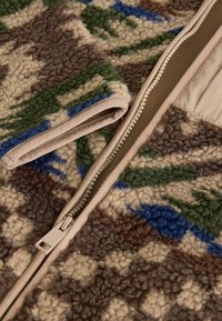 Fleece jacket featuring a camouflage pattern in brown, green, and blue. Includes a zipper with a beige fabric trim. Soft texture throughout.