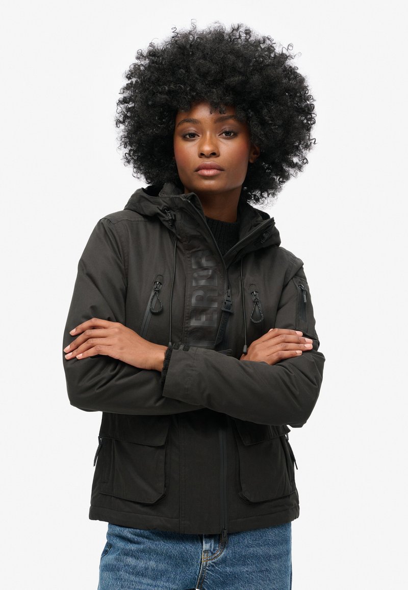 Schwarze Softshelljacke mit Kapuze, ausgestattet mit elastischen Kordeln, Reißverschluss- und Klappentaschen sowie einem Logodetail auf der Vorderseite.