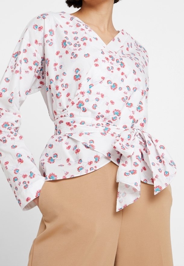 Femme portant un chemisier cache-cœur blanc à fleurs bleues et rouges, noué à la taille, associé à un pantalon marron clair.