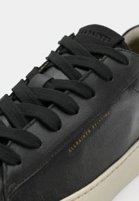 Sneaker in pelle nera con lacci neri piatti, logo "ALLSAINTS" in oro embossato e suola in gomma color crema, con una texture liscia.